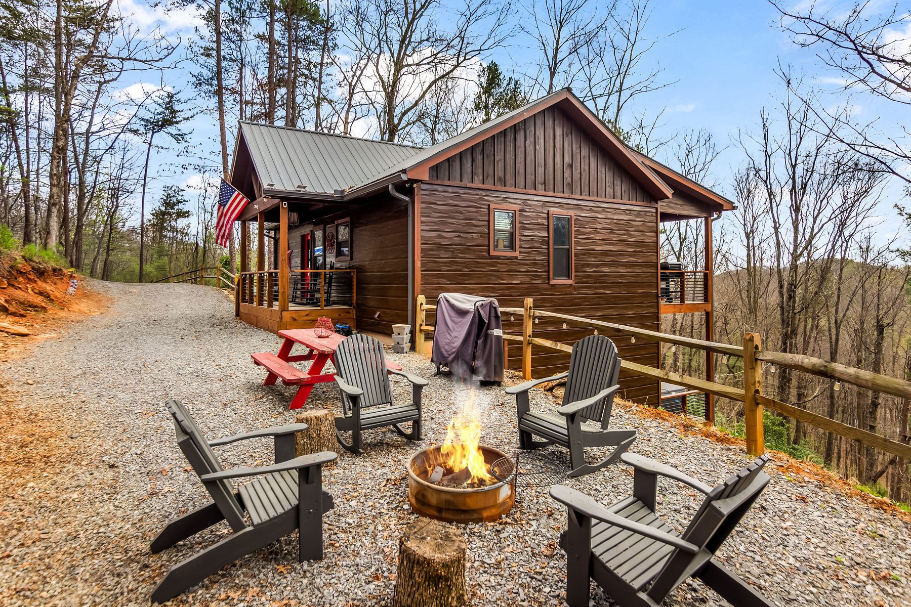 Cabin exterior with fire pit and Adirondack chairs in the foreground, gravel driveway curving past