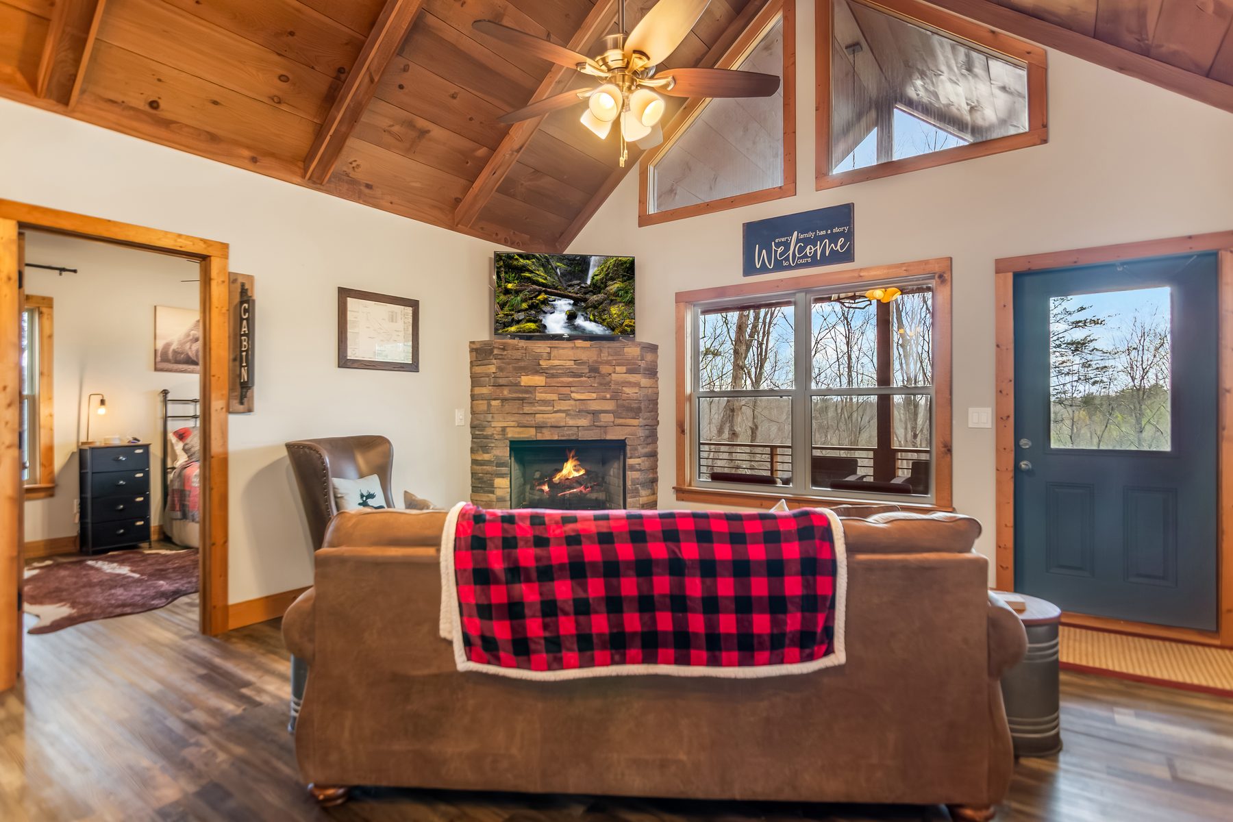 Stone fireplace with the fire lit, leather sofa with plaid throw, looking out to the woodland deck