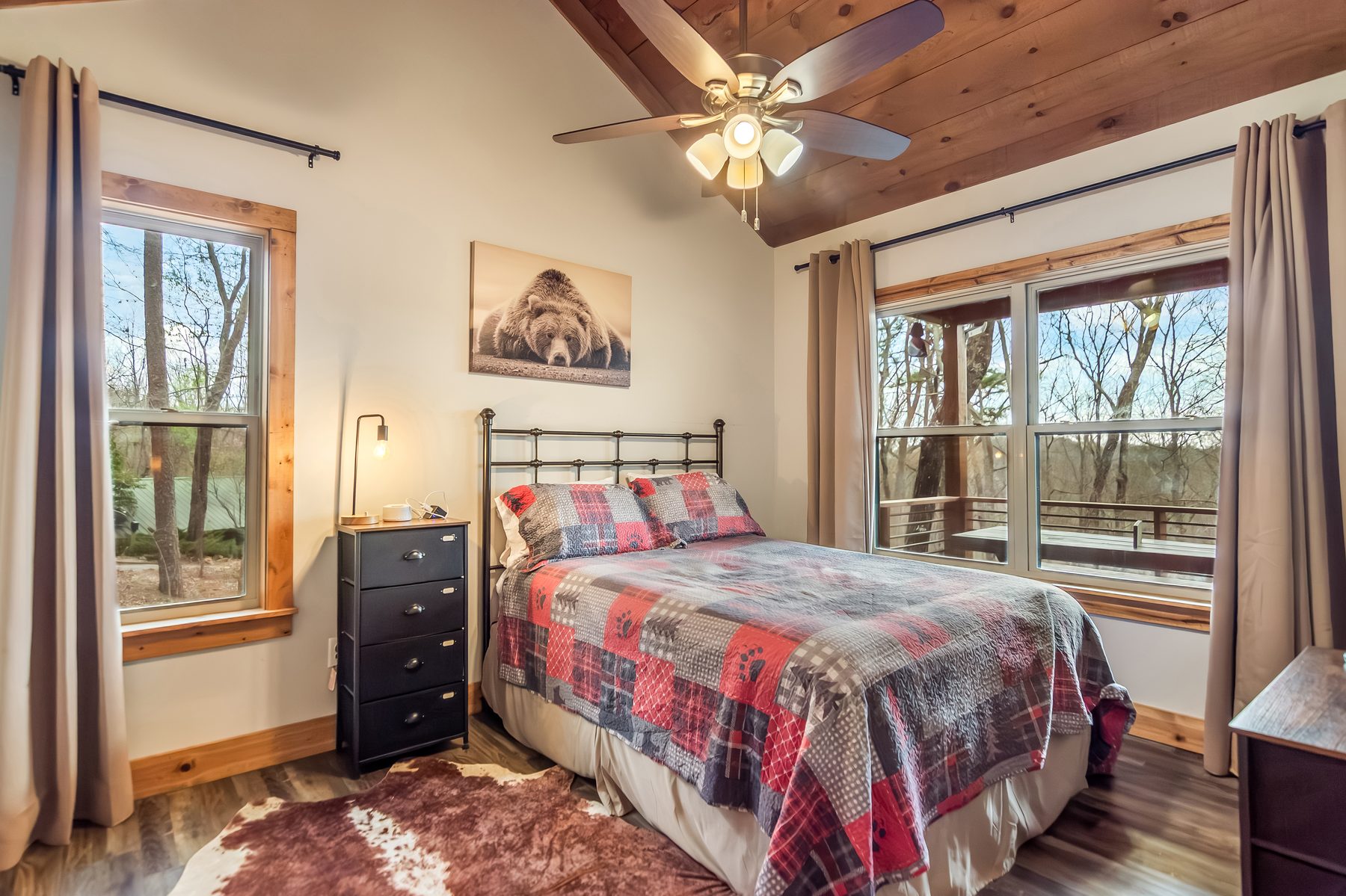 Queen bedroom with iron headboard, plaid bear quilt, bear photo print, and woodland window views