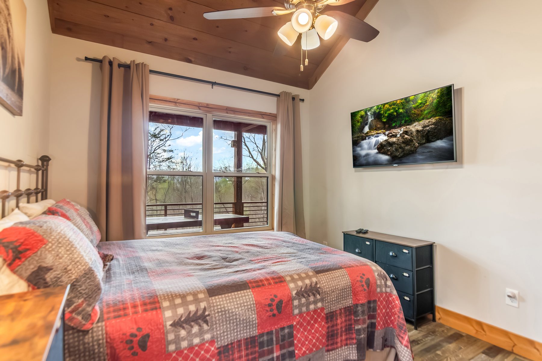 Queen bedroom with bear-themed plaid quilt, mounted TV, and sliding door to the deck