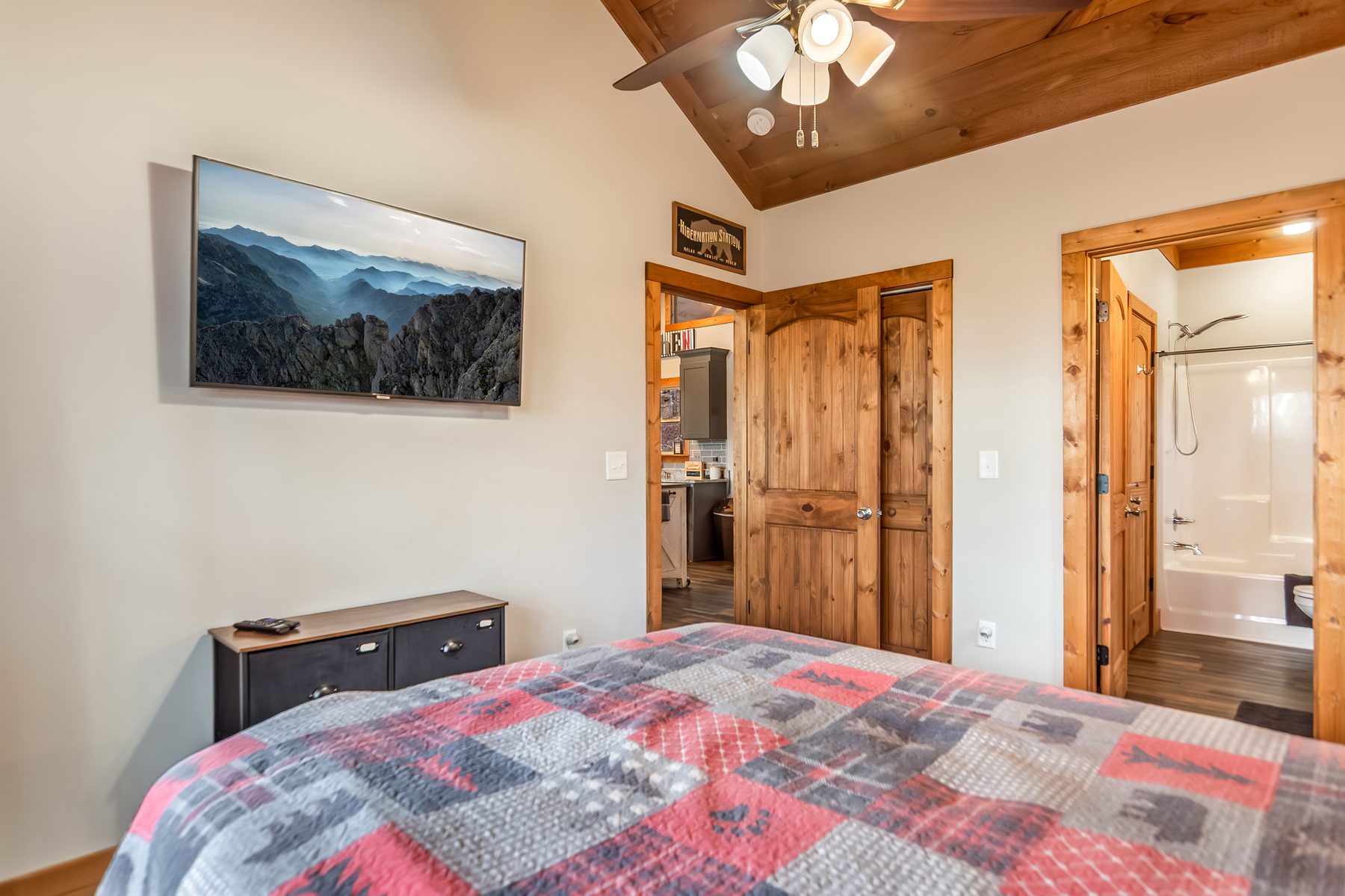 Bedroom with mountain-landscape print over plaid quilt and doorway to the en-suite bath
