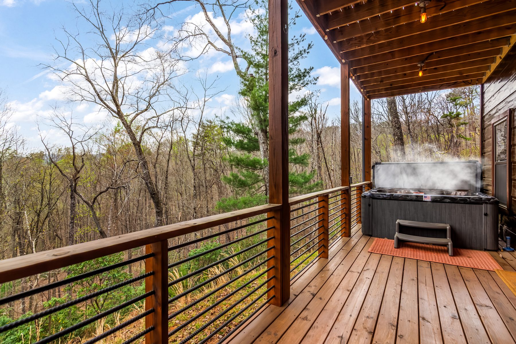 Covered porch with cable railings, hot tub at the far end, and woodland views